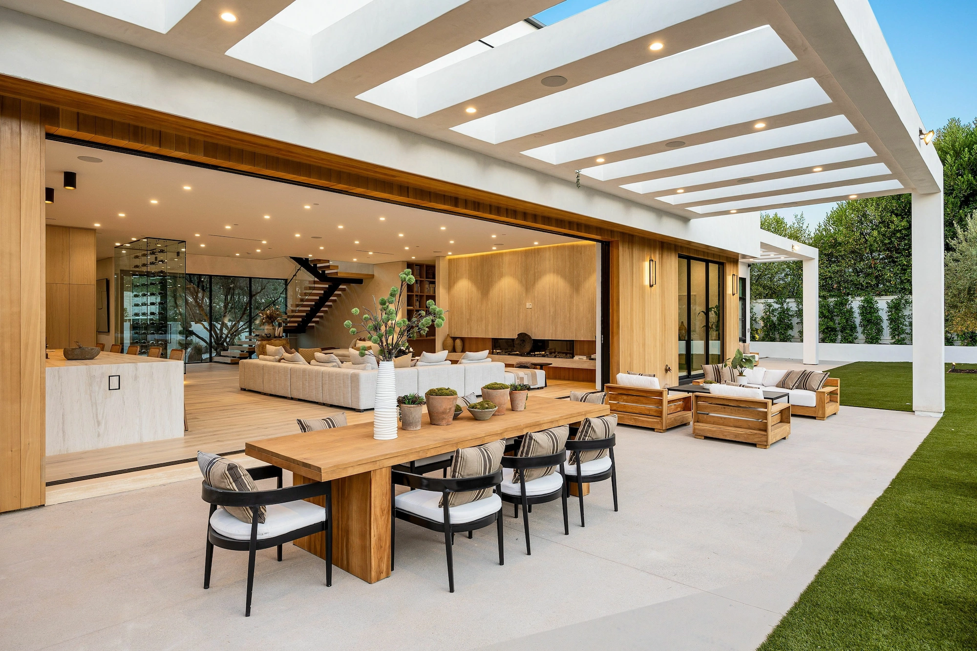 A modern outdoor living space featuring a wooden dining table with eight chairs, surrounded by potted plants. The background shows a spacious interior with a comfortable sofa and a staircase, visible through a large opening that connects the outdoor and indoor areas. Natural light floods the space through the skylight and large glass walls.